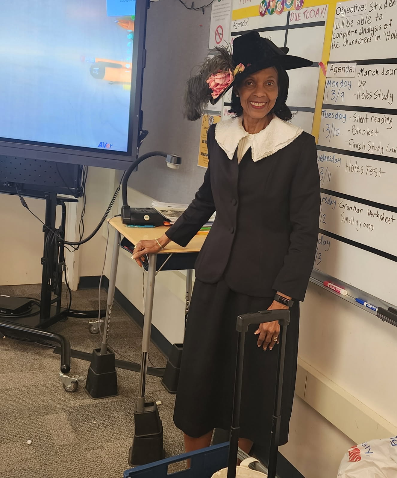An African American woman smiles while dressed in a period costume in a school classroom. She is depicting Madame C.J. Walker, outfitted in a long black suit with a lace white collar and a wide-brimmed black hat.