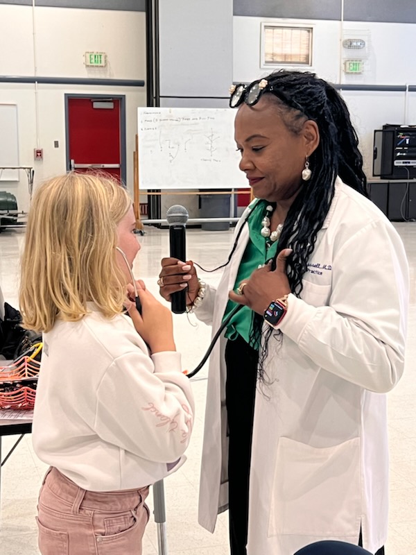 A physician in a white lab coat stands before a child who listens to the doctor's heart using a stethoscope.
