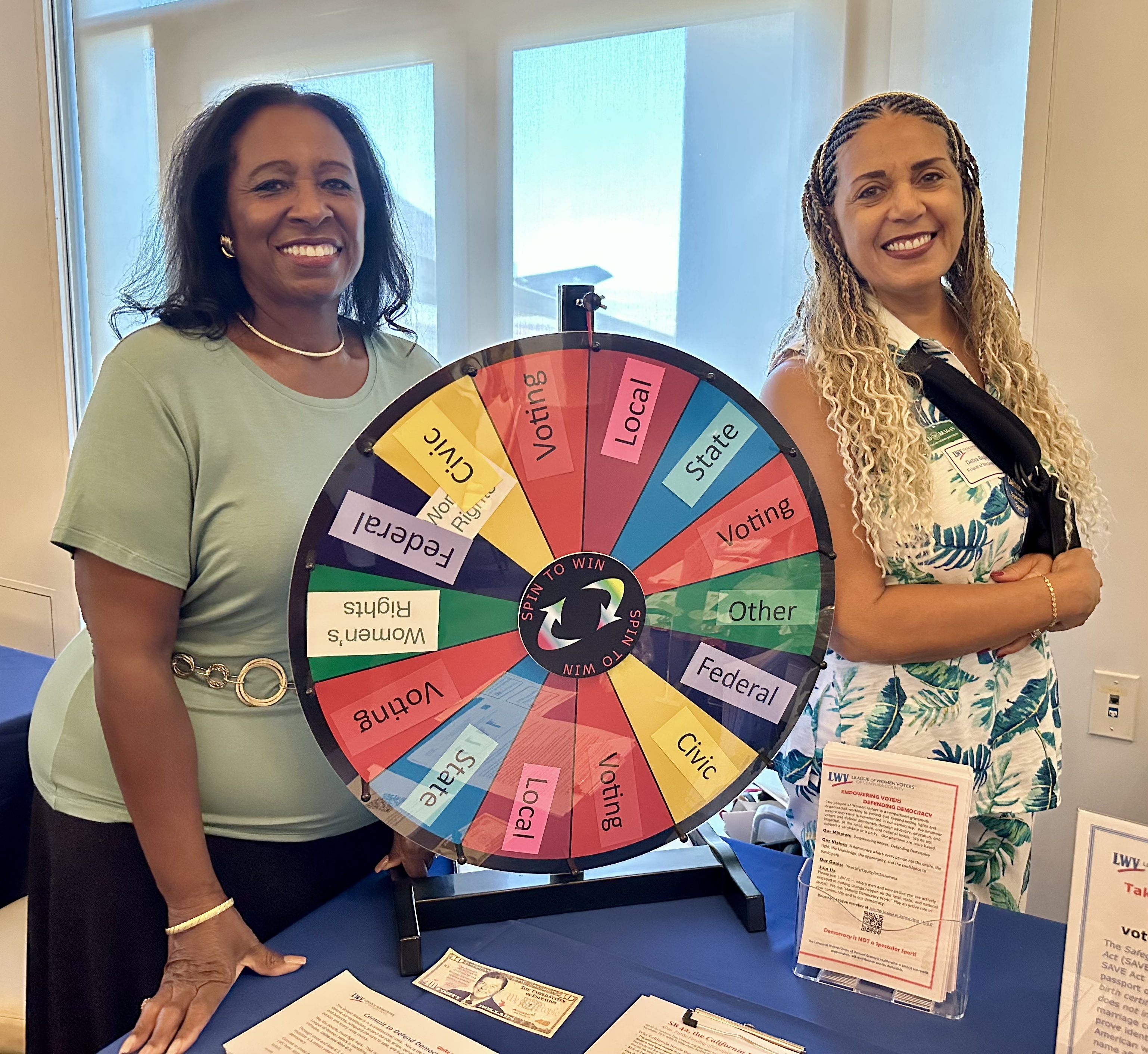 A colorful spin wheel with words relating to voting in the United States is at the center of the frame and two nicely dressed African American women flank the image.