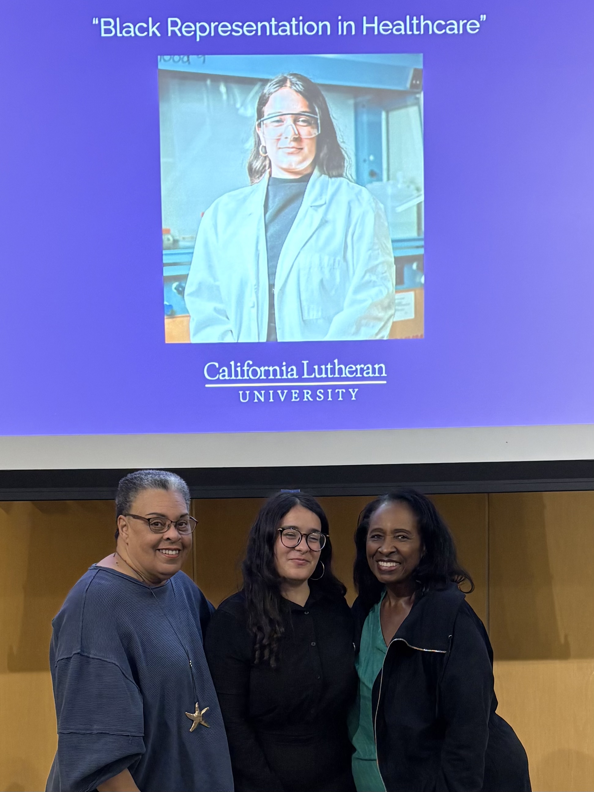 Two chapter members flank a college student. The three are before a powerpoint slide showing the student in a white lab coat.