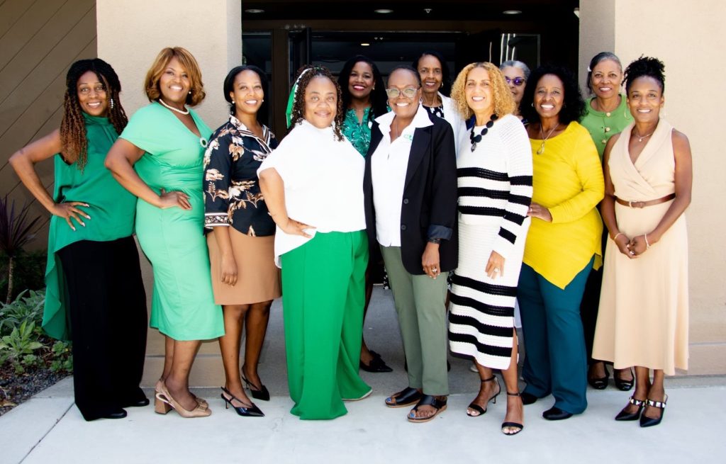 Twelve African-American women are clustered near a hotel entrance, smiling. Many are dressed in shades of green and white.
