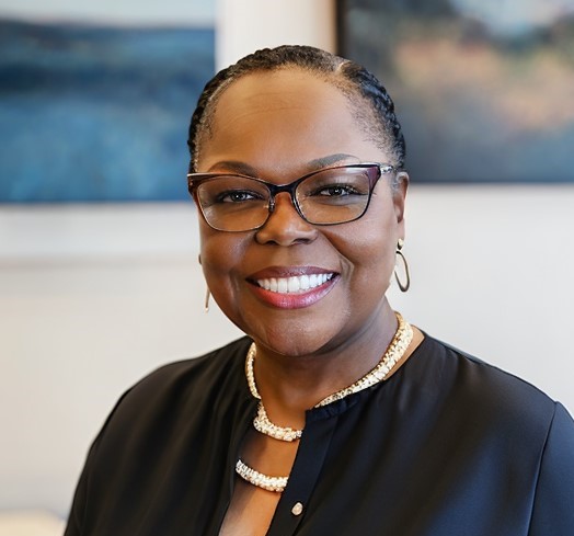 Brown-skinned woman wearing glasses, black top, and gold jewelry smiles in a professional photo